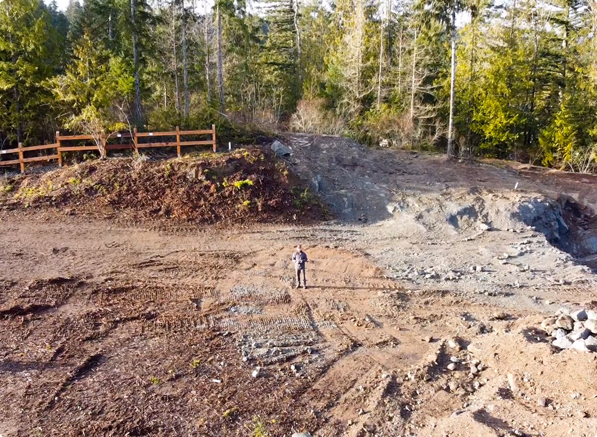 A person standing on a cleared dirt area with tire tracks, surrounded by forest and a wooden fence in the background.