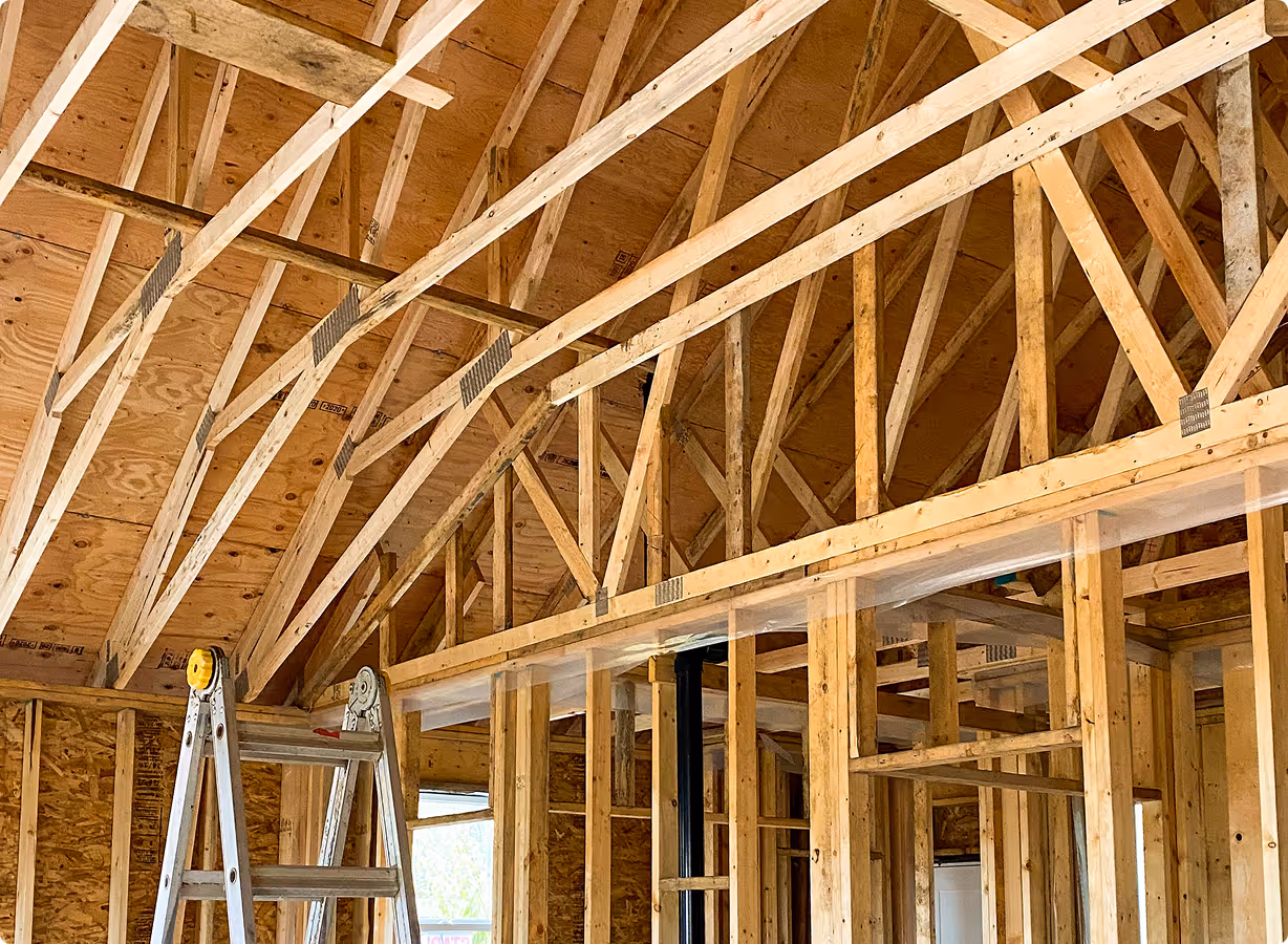Interior framing of a wooden roof structure with exposed trusses and a metal folding ladder inside an unfinished building.