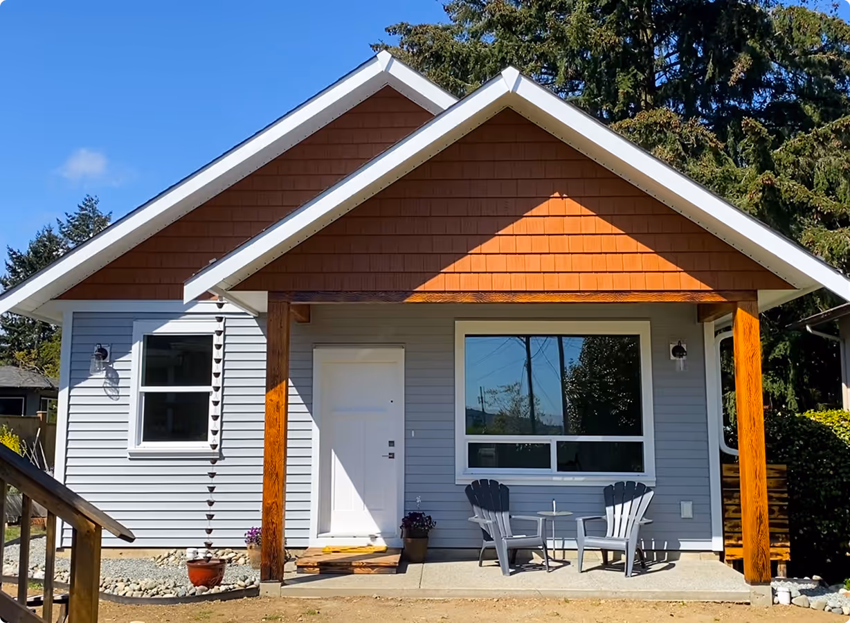 Small modern house with blue siding, brown gable roof, white door, and a porch with two gray chairs and a small table.