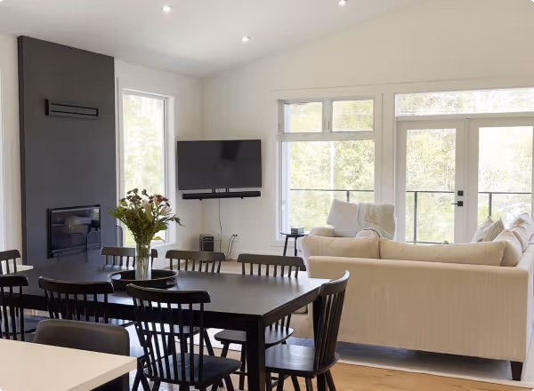 Bright living room with a beige sectional sofa, black dining table with chairs, wall-mounted TV, and large windows overlooking greenery.