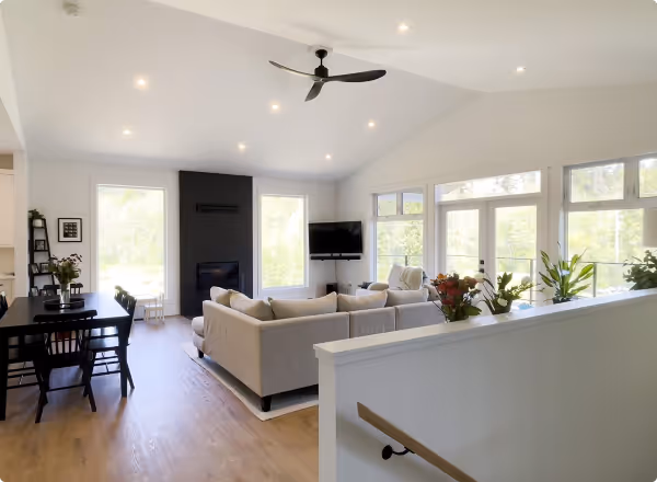 Spacious open living room with beige sofas, a black fireplace, wall-mounted TV, dining table, ceiling fan, and large windows letting in natural light.
