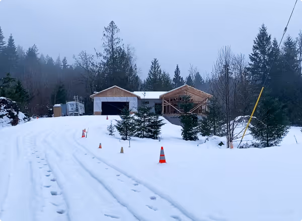 Snow-covered driveway leading to a partially built house surrounded by evergreen trees on a cloudy day.