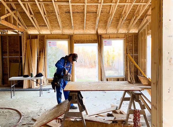 Construction worker in blue coveralls working inside a house under construction, surrounded by wooden framing and plywood sheets.
