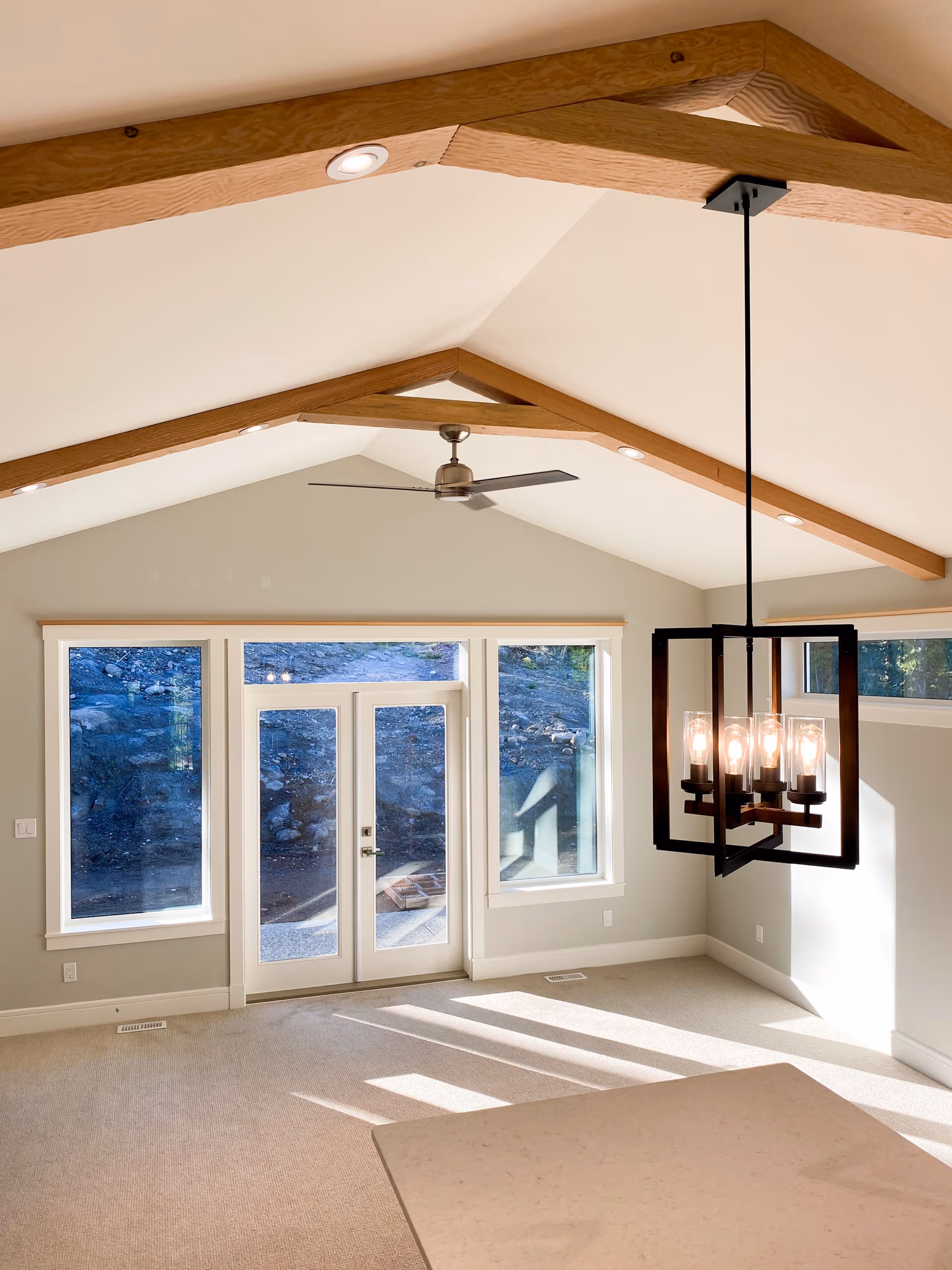 Bright room with beige carpet, large windows and glass door revealing rocky outdoor area, wooden ceiling beams, ceiling fan, and modern black pendant light fixture.