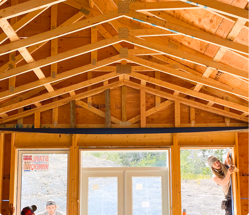 Interior of a wooden framed house under construction with exposed ceiling beams and workers installing a window.