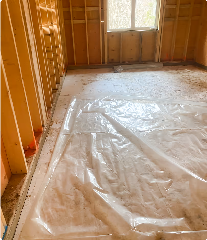 Interior of a room under construction with exposed wooden framing and a plastic sheet on the floor beneath a large window.