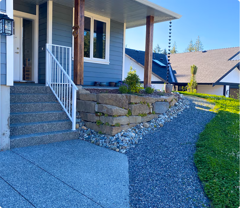 Front porch of a house with gray steps, stone retaining wall, gravel pathway, and landscaped green lawn under a clear blue sky.