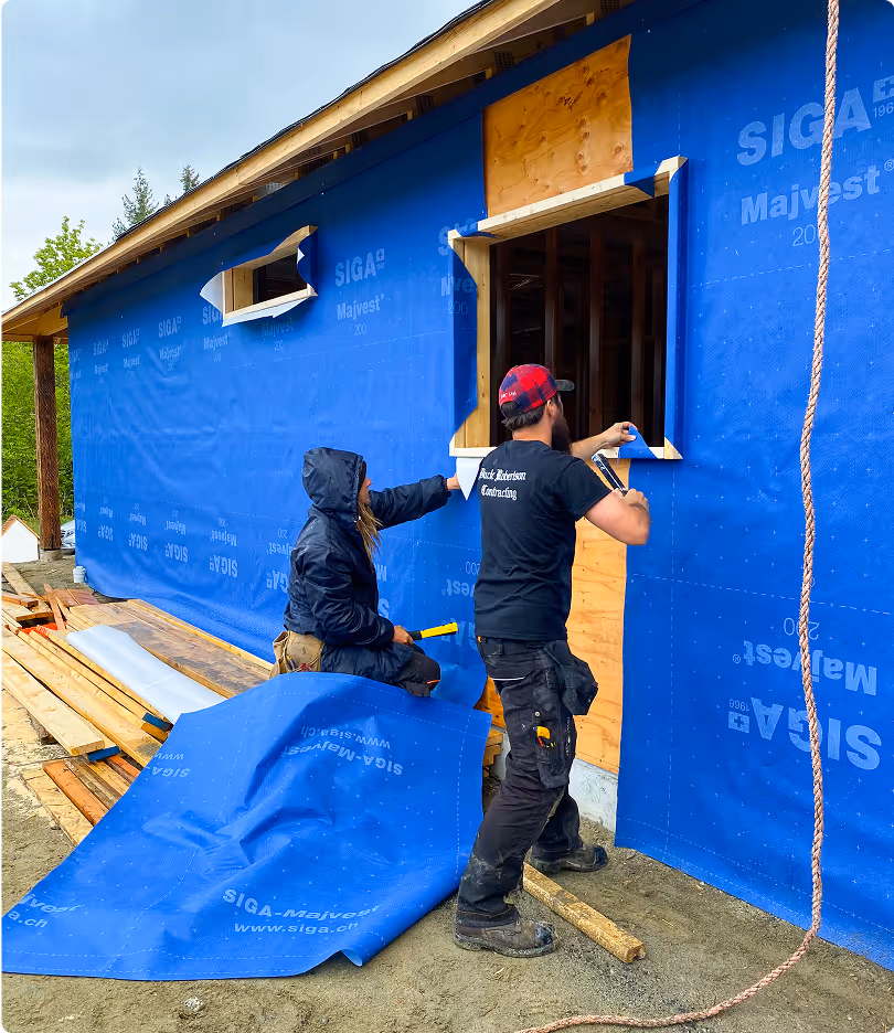 Two construction workers installing blue waterproof house wrap around window openings on a wooden-frame building.