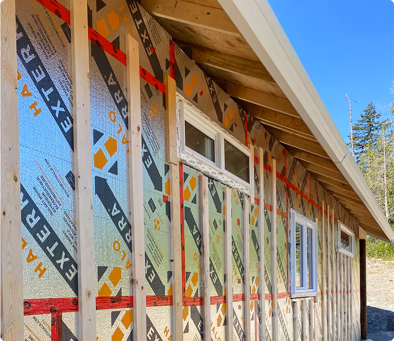 Side view of a wooden building under construction with reflective weather barrier and three windows, set against a clear blue sky and trees.