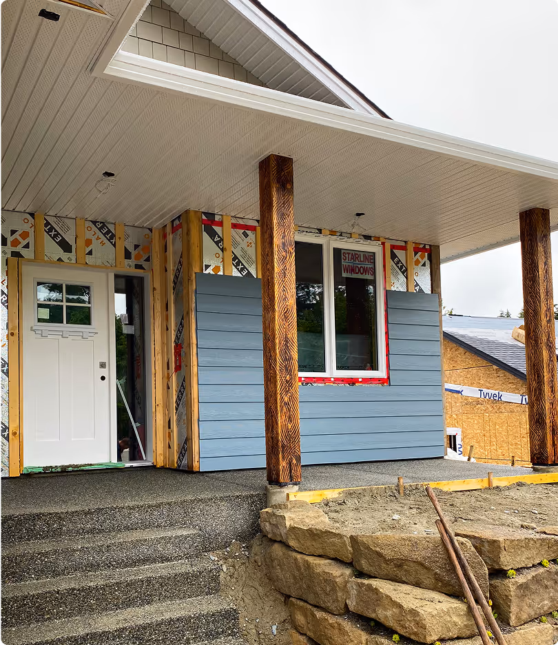 Under-construction house porch with blue siding partially installed, white front door, wooden columns, and stone retaining wall.