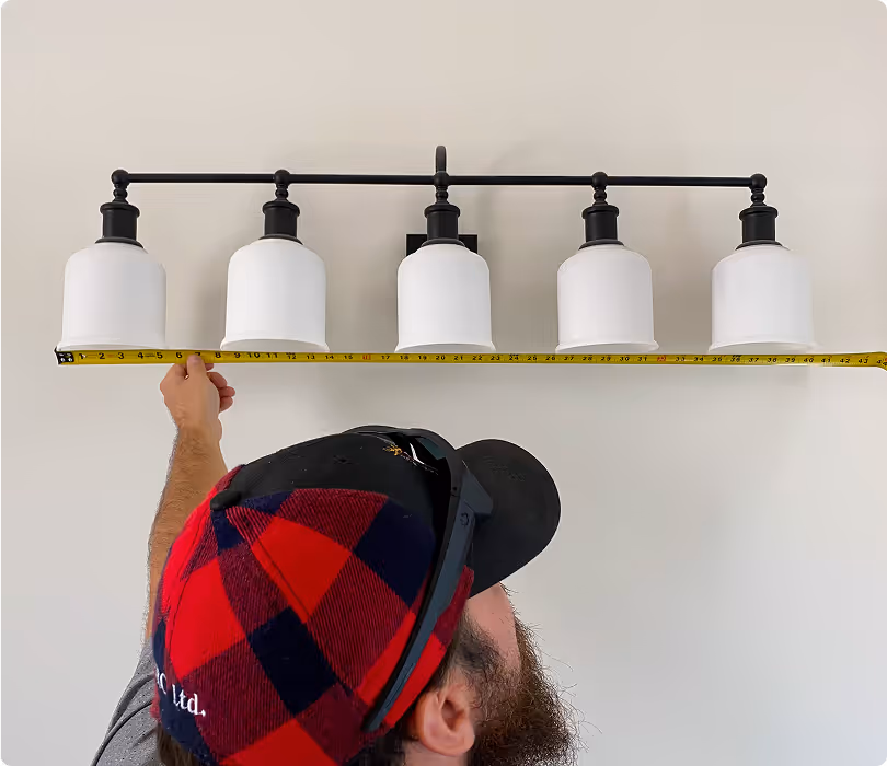 Man in a red and black checkered cap measuring the width of a five-light bathroom vanity fixture with a yellow tape measure.