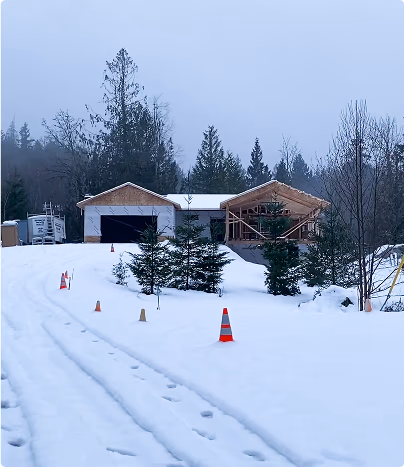 Snow-covered driveway with footprints leading to a partially constructed house surrounded by trees in a foggy setting.