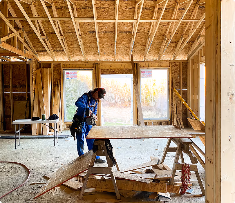 Construction worker in blue coveralls working with a power tool on a plywood sheet inside a partially built wooden house.