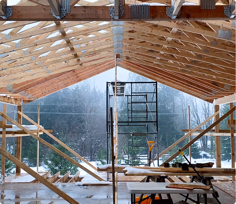Interior view of a wooden building under construction with exposed trusses and scaffolding, snow visible outside.