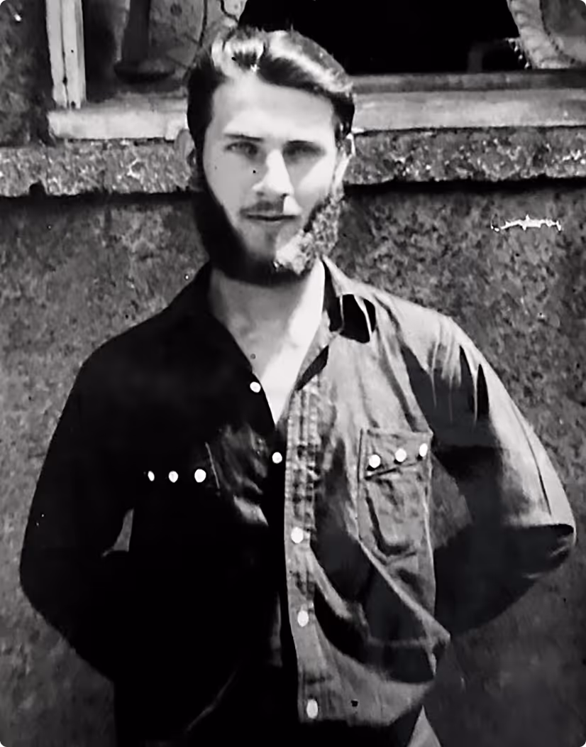 Black and white photograph of a young man with a beard wearing a buttoned shirt standing in front of a textured wall.