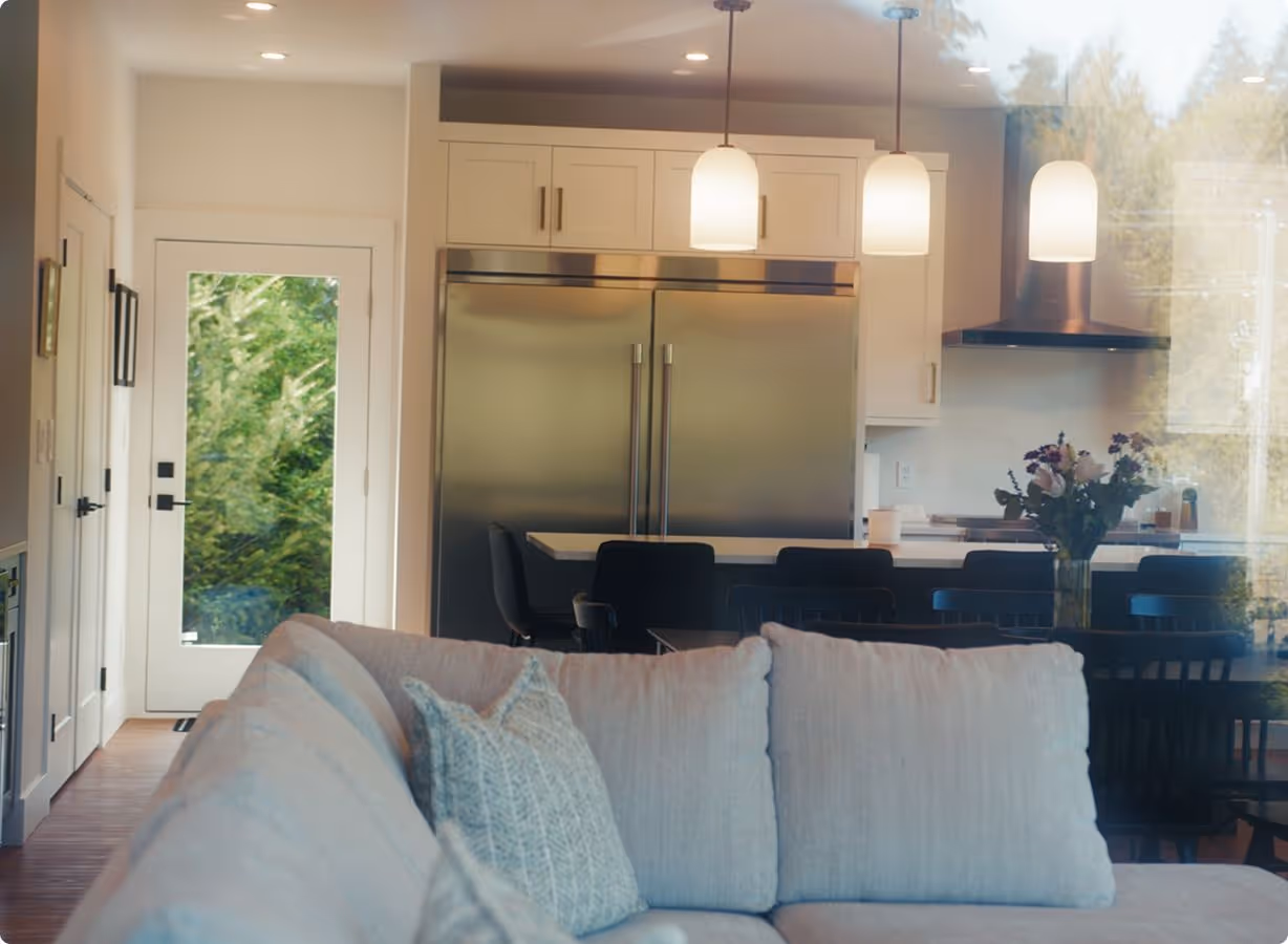Modern kitchen with stainless steel double-door refrigerator, white cabinets, hanging pendant lights, and a dining table with a flower vase, viewed through a glass window with a gray couch in the foreground.