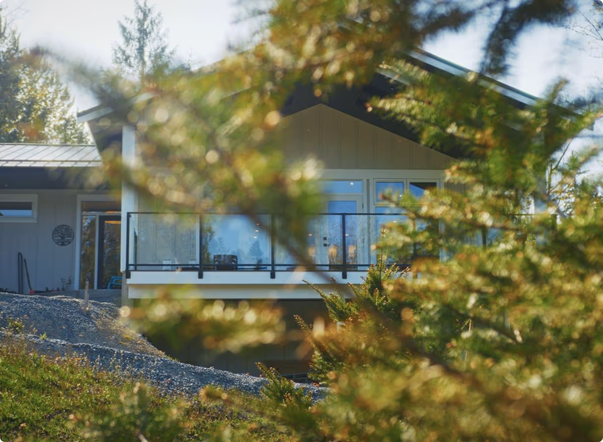 View of a modern house with a glass balcony, partially obscured by evergreen tree branches in the foreground.