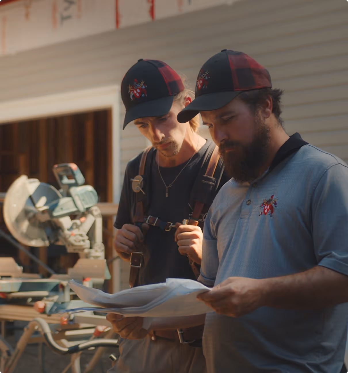 Two construction workers in matching hats examine a set of building plans at a worksite with tools in the background.