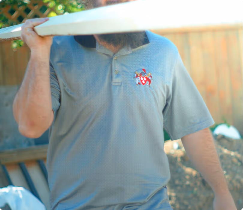 Man in a grey polo shirt carrying a wooden plank on his shoulder outdoors.