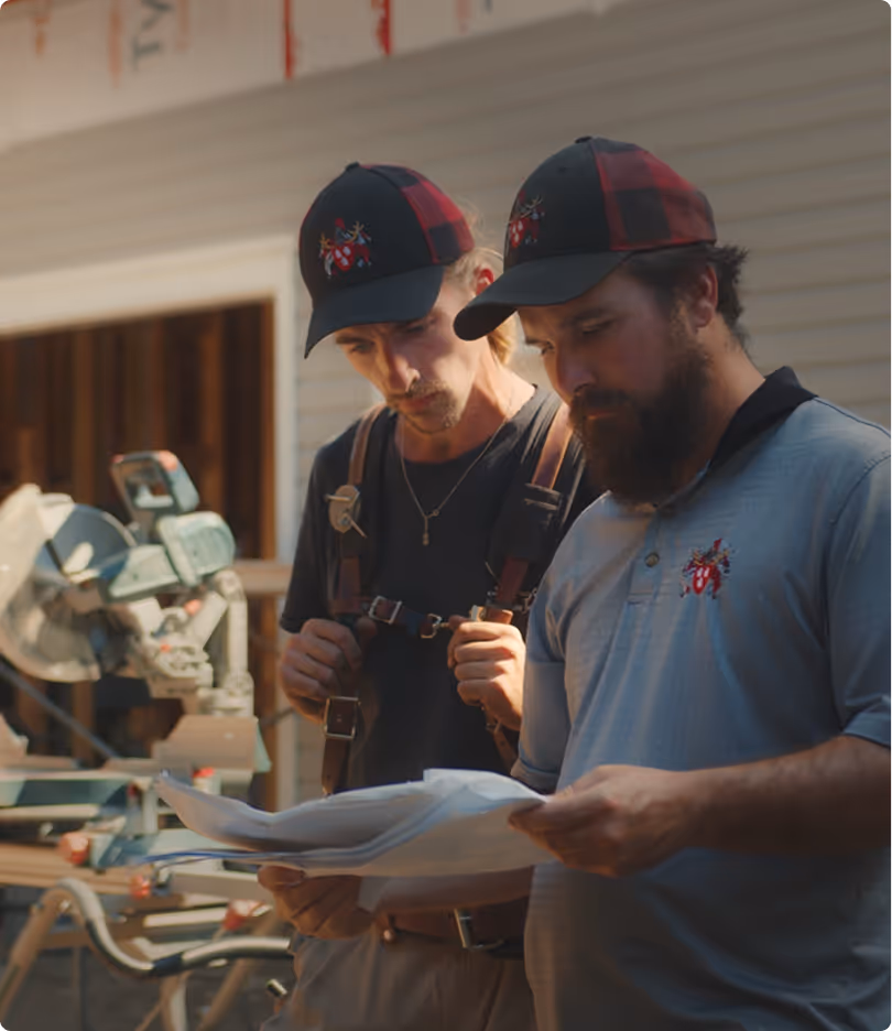Two construction workers wearing matching black and red hats reviewing architectural plans on a job site.
