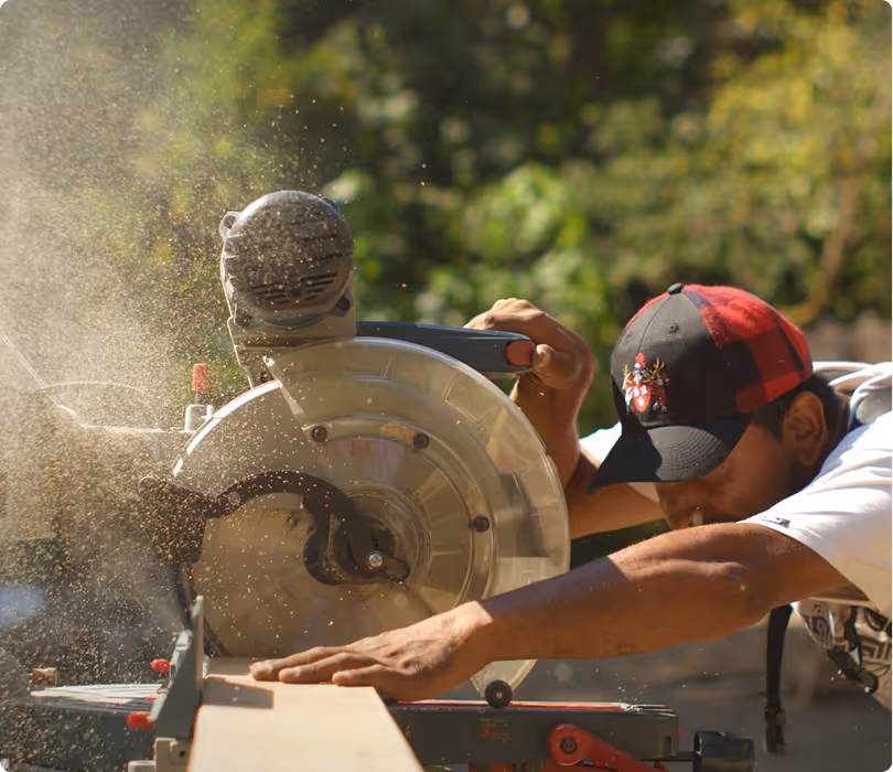 Man wearing a red and black cap using a circular saw to cut a wooden board outdoors with sawdust flying.