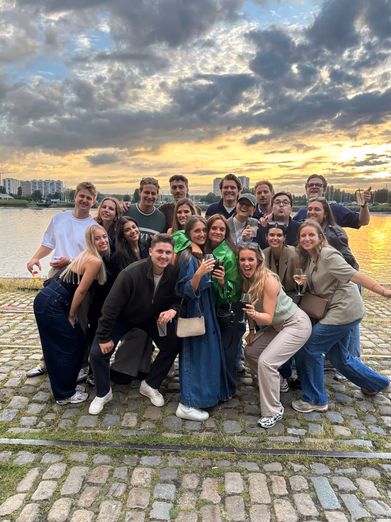Group of young adults posing happily on cobblestone ground by a river during a sunset with cloudy sky.