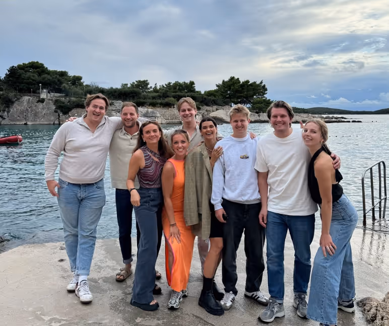 Group of nine young adults standing close together and smiling on a concrete dock by a calm body of water with rocky shore and trees in the background under a cloudy sky.