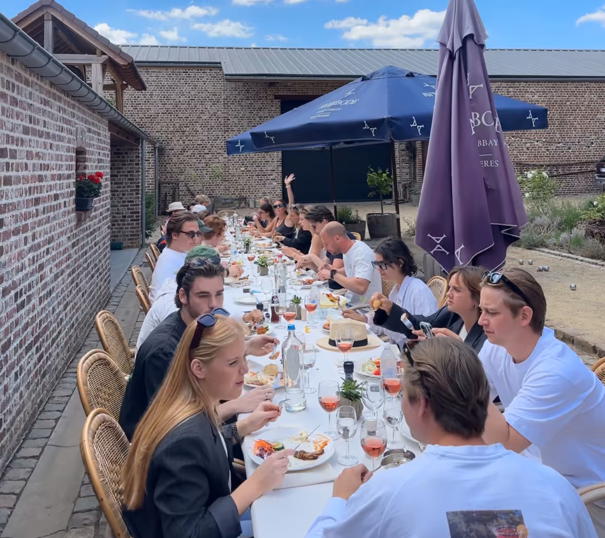 Group of people enjoying an outdoor meal at a long table set with plates, wine glasses, and bottles under blue and purple umbrellas.