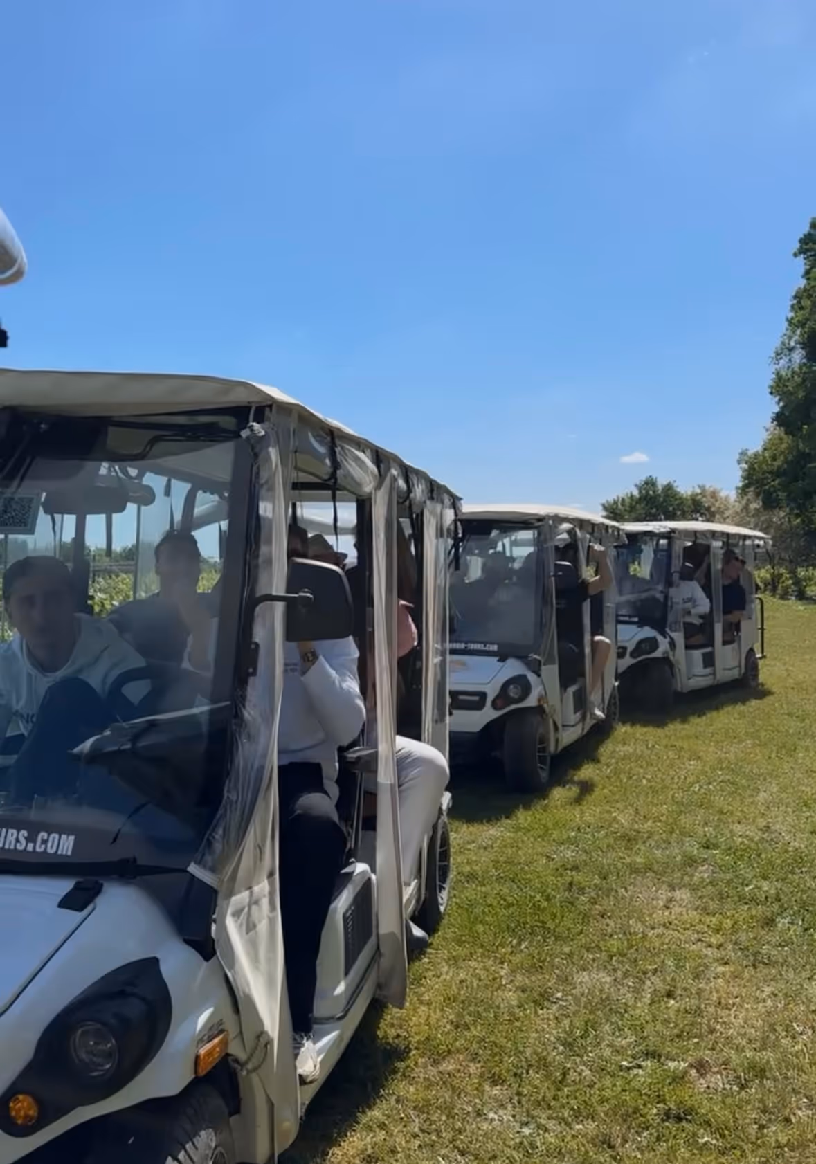 Three white golf carts with clear plastic sides are parked in a row on grass under a blue sky, each carrying multiple passengers.