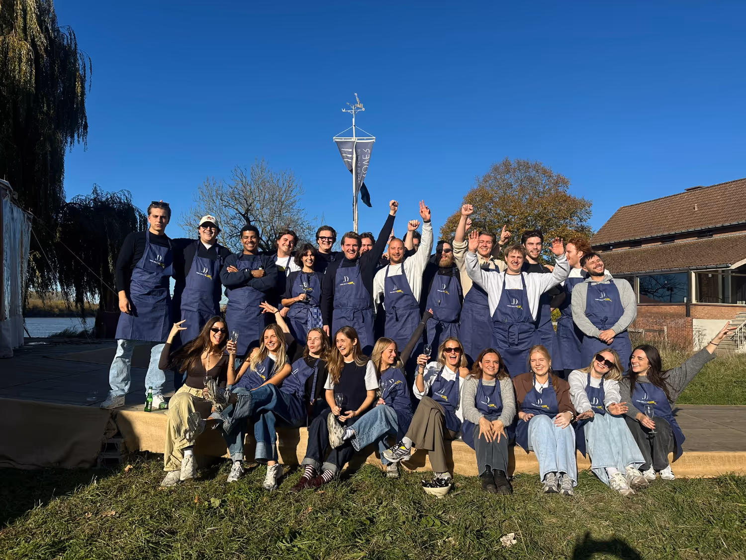 Group of people outdoors on a sunny day wearing navy aprons, smiling and posing cheerfully on grass and a stage.