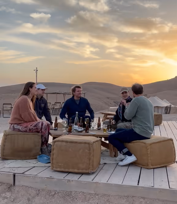 Five people sitting on cube cushions around a low wooden table outdoors at sunset in a desert setting.