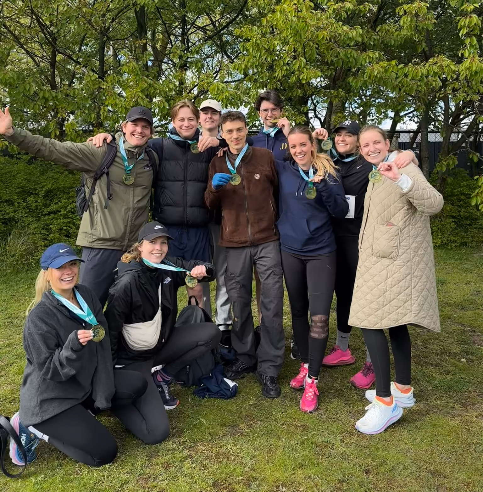 Group of nine smiling young people outdoors showing medals, gathered on grass with green trees in the background.
