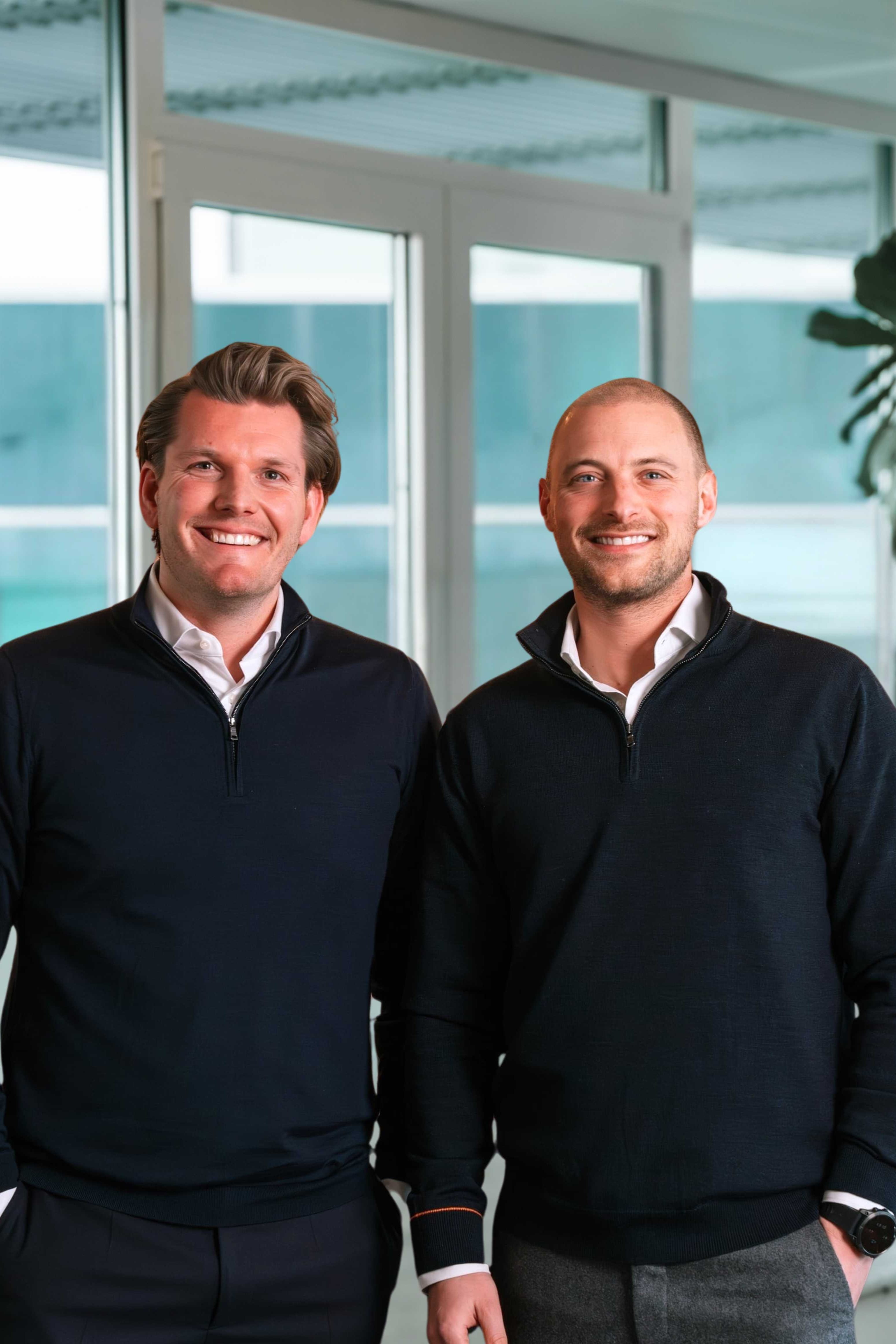 Two smiling men in casual business attire posing by a pool table in a bright room with large windows.