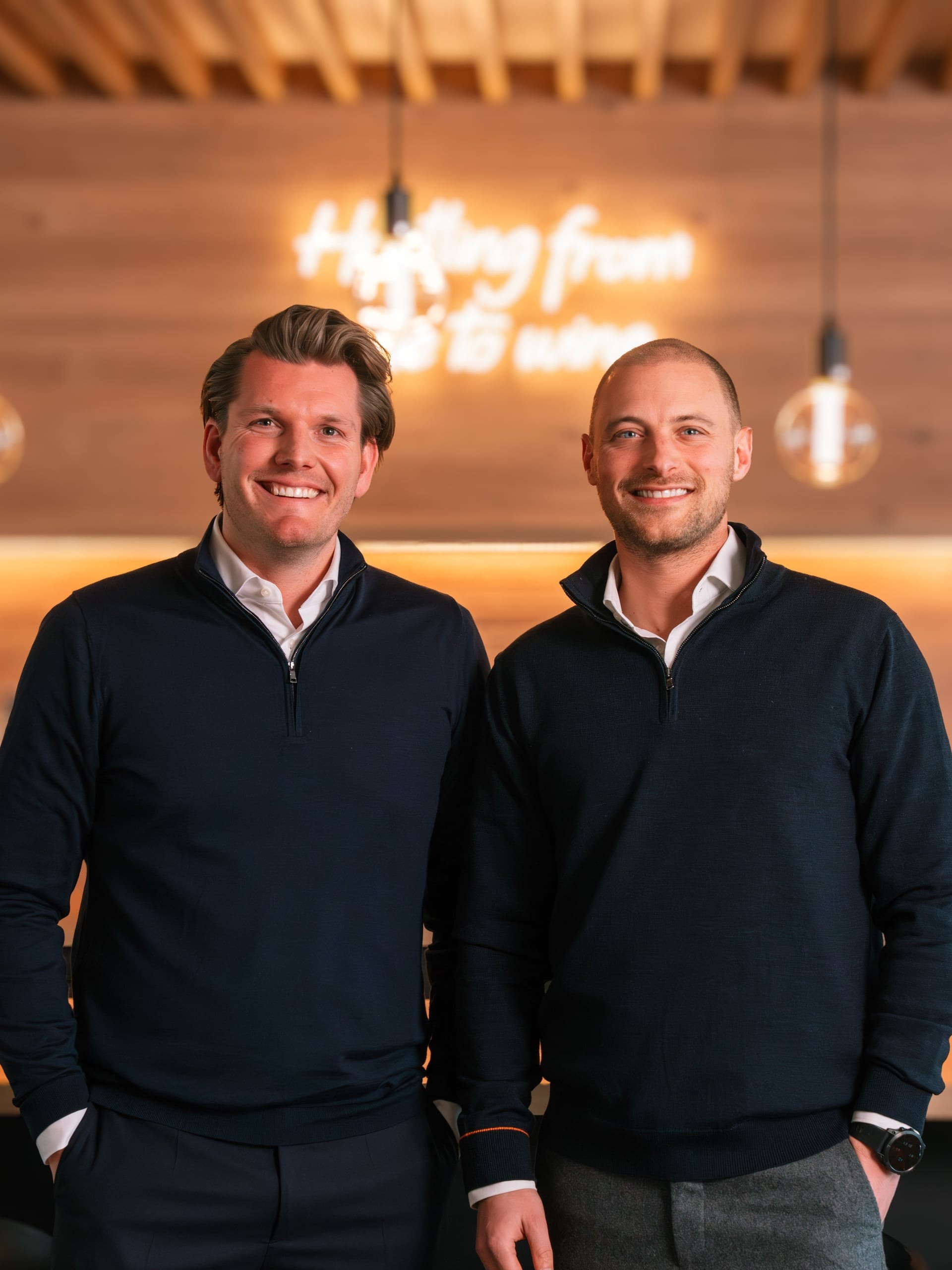 Two smiling men in casual business attire posing by a pool table in a bright room with large windows.
