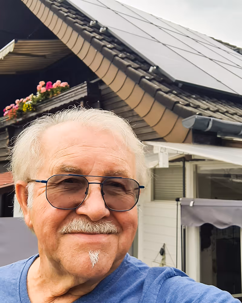 Älterer Mann mit Sonnenbrille lächelt vor seinem Haus mit Solaranlage