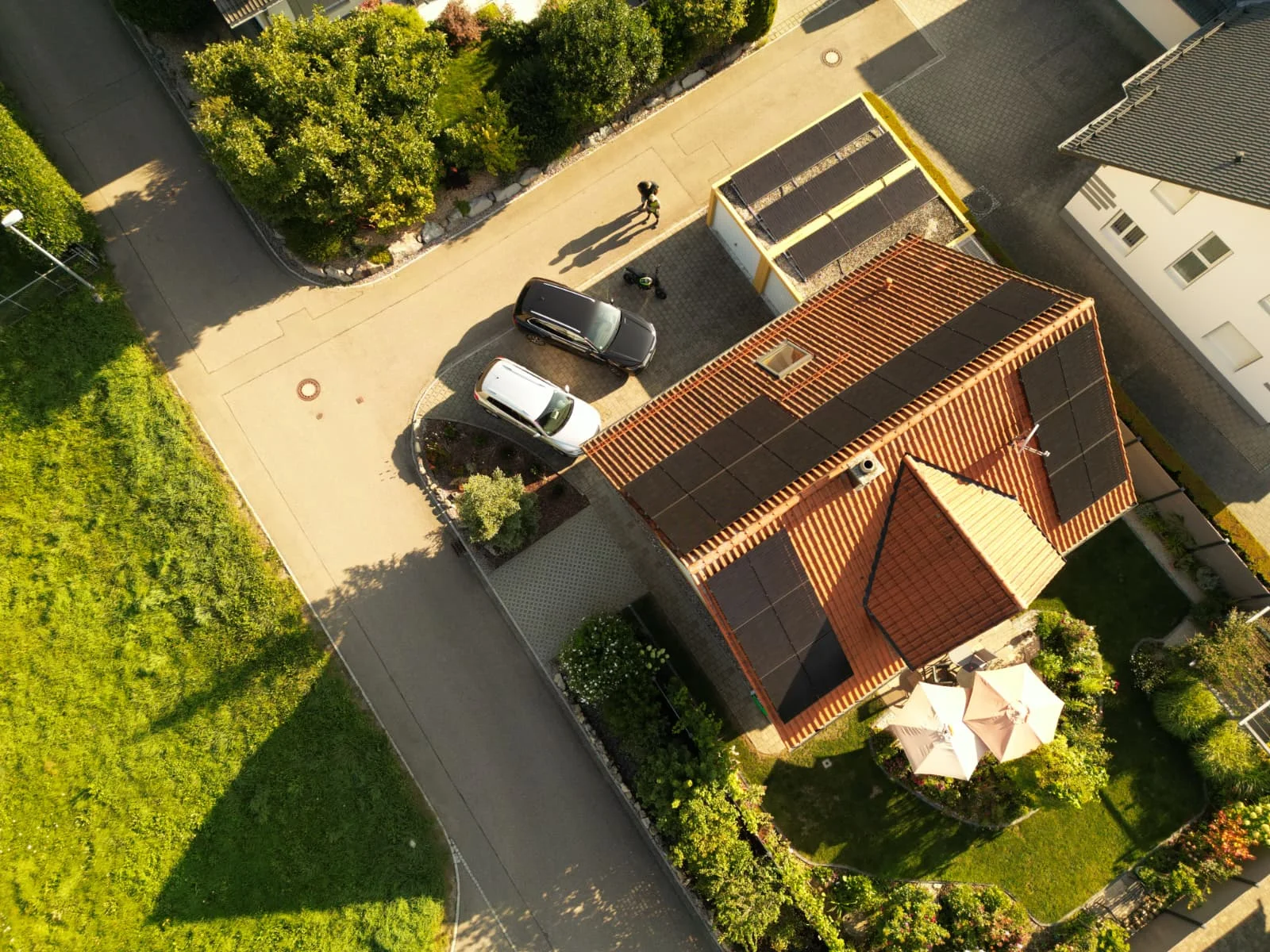 Vogelperspektive auf ein Haus mit Solarpanels auf dem Dach, zwei geparkte Autos und eine Terrasse mit Sonnenschirmen im Garten.