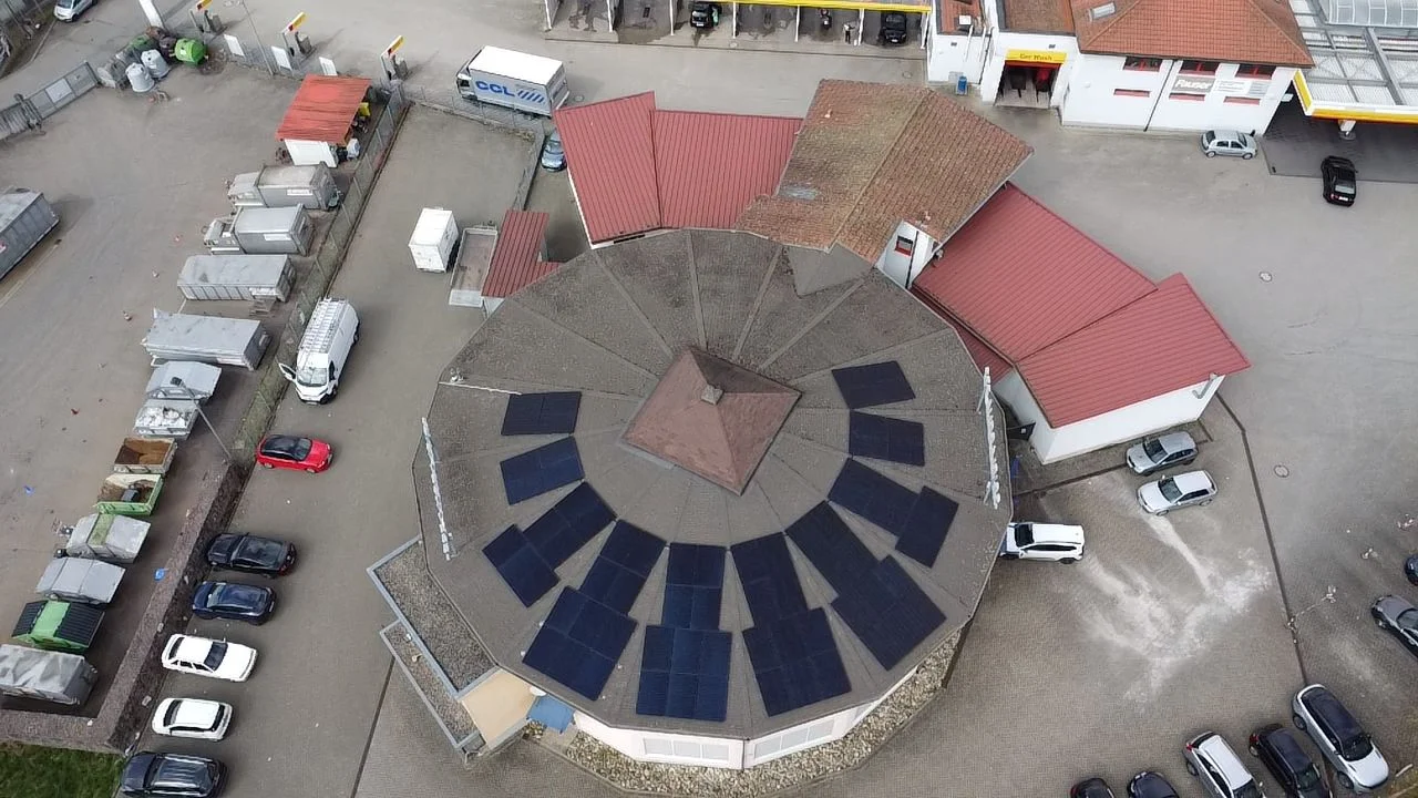 Luftaufnahme eines runden Gebäudes mit auf dem Dach installierten Solarzellen, umgeben von geparkten Autos und Containern.