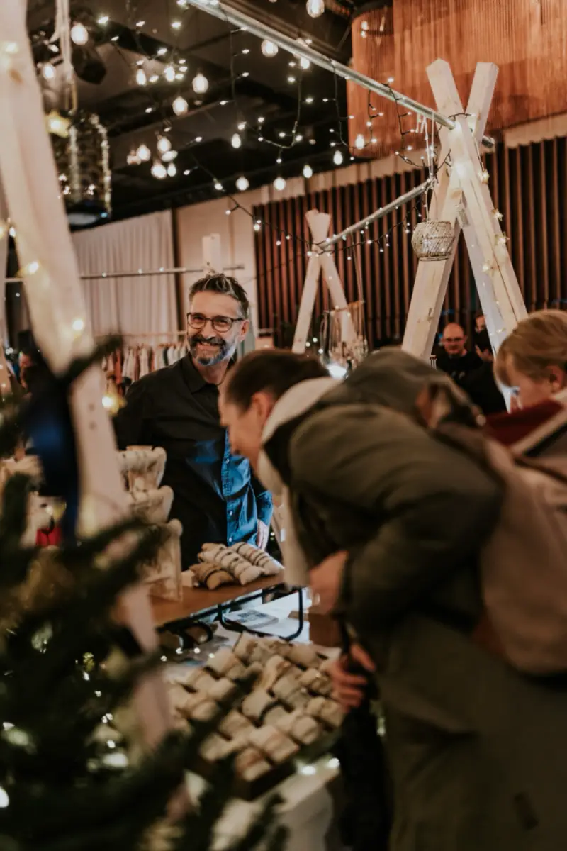 Lächelnder Verkäufer auf einem Weihnachtsmarkt, umgeben von Kunden und Lichtern