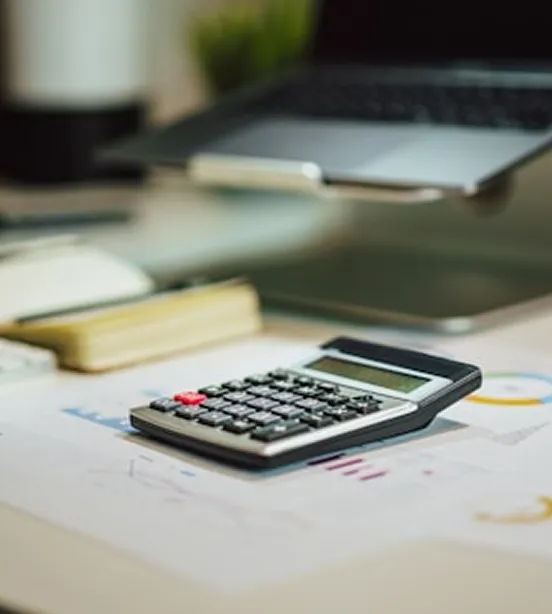 Calculator on desk with charts and laptop.