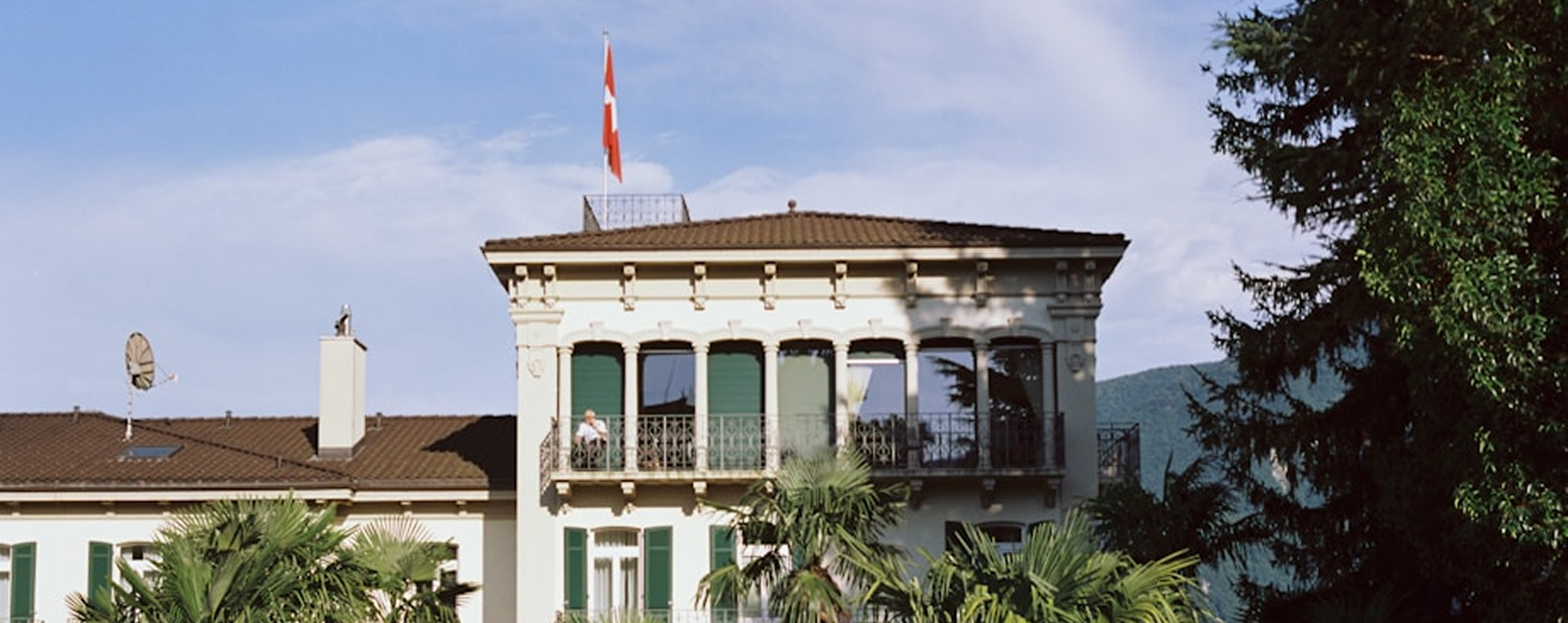 Elegant cream-colored building with green shutters and balconies, topped with a Swiss flag, surrounded by palm trees under a blue sky.