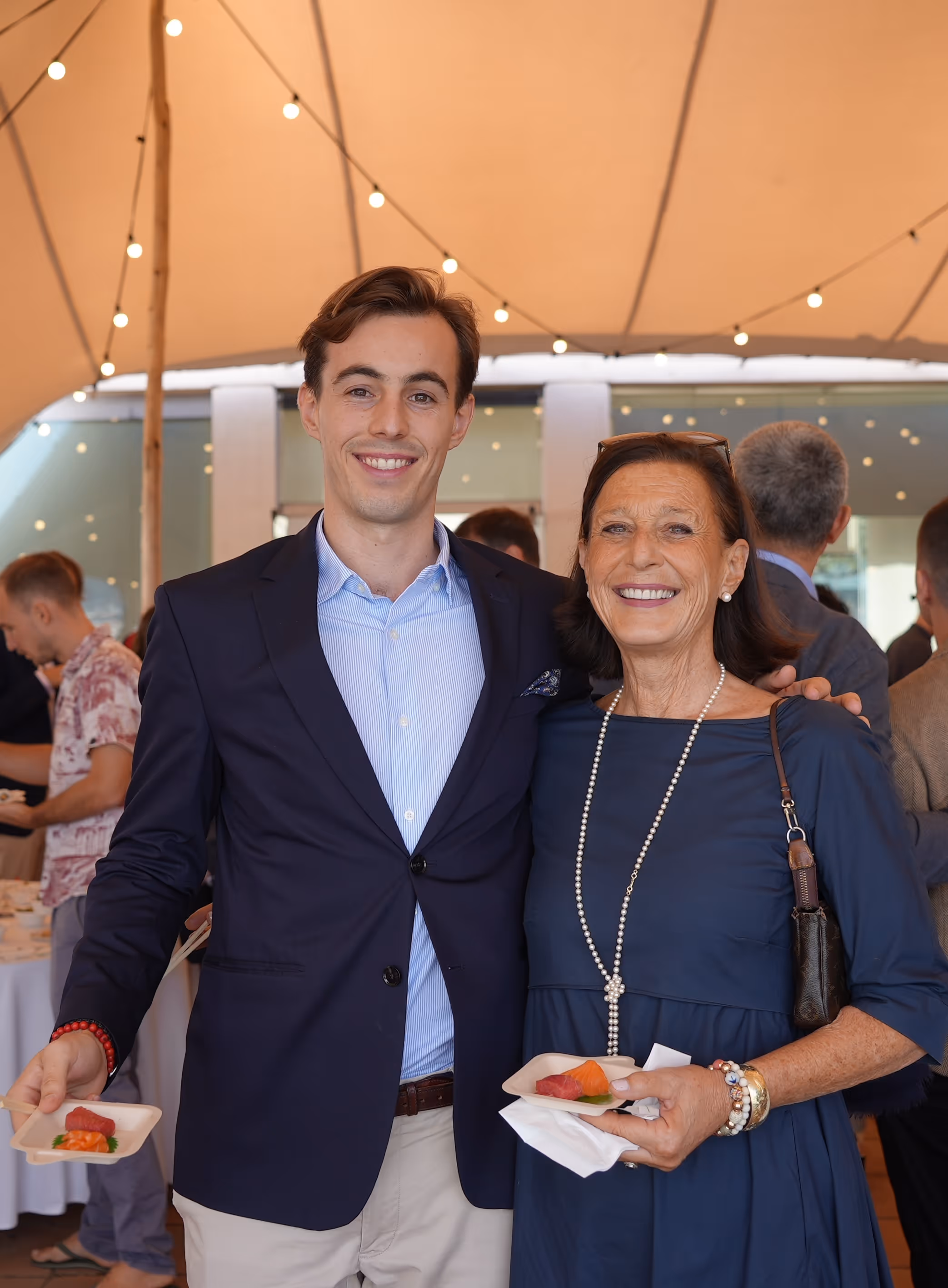 Smiling man called Moritz von Oheimn and woman called Eva von Oehimb in a blue dress holding plates of sushi at an outdoor social event under string lights.