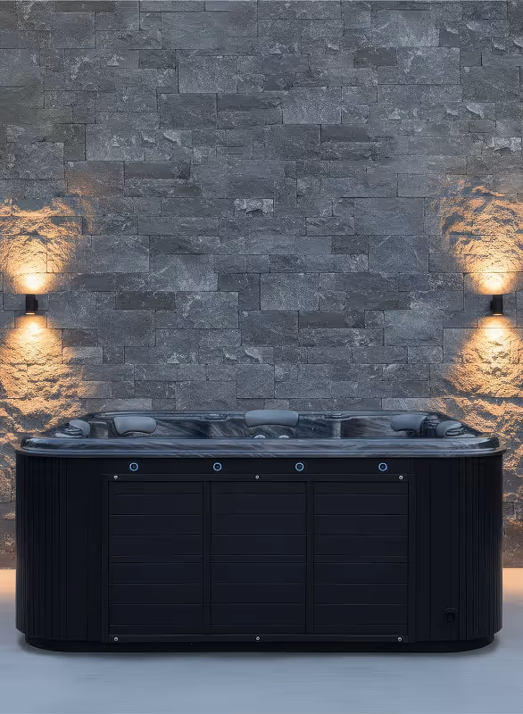 Modern black hot tub with built-in headrests against a textured gray stone wall lit by two wall sconces.