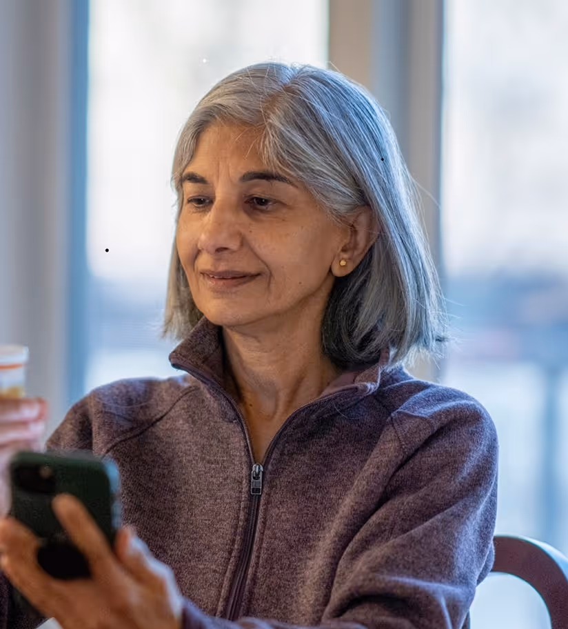 Middle-aged woman with gray hair holding a prescription pill bottle and a smartphone, sitting indoors near a window.