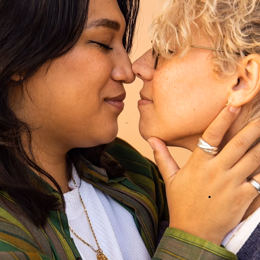 Two people facing each other closely, one with dark hair and the other with curly blonde hair and glasses, sharing an intimate moment.