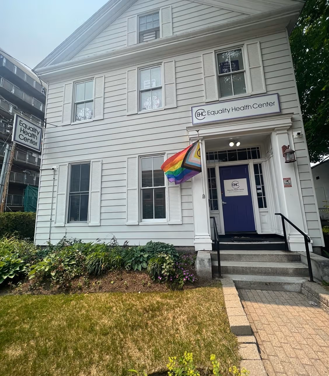 White two-story building with a purple door displaying a rainbow Progress Pride flag and signs reading Equality Health Center.