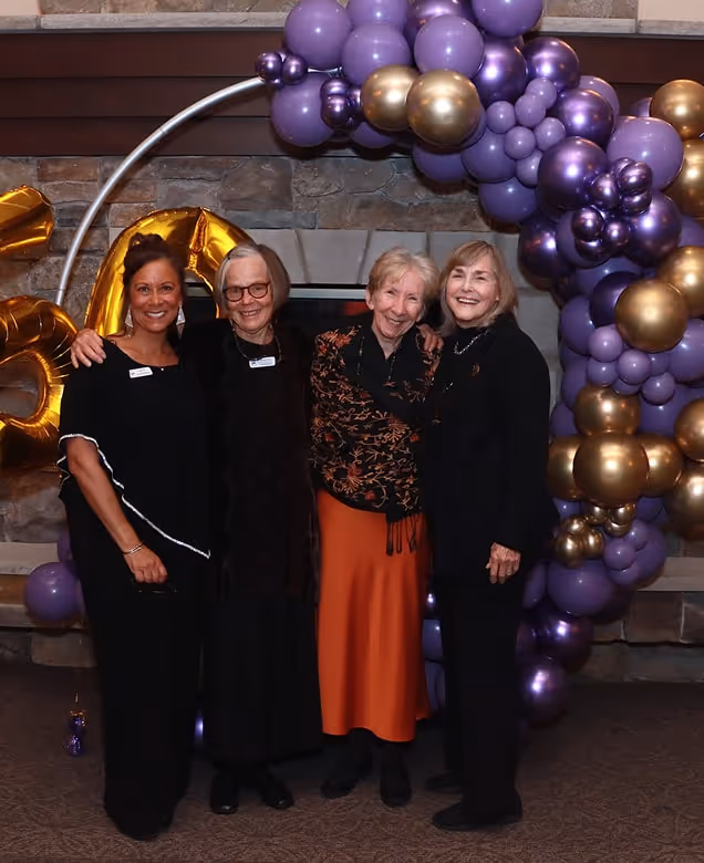 Four smiling women standing together in front of a stone fireplace decorated with purple and gold balloons.