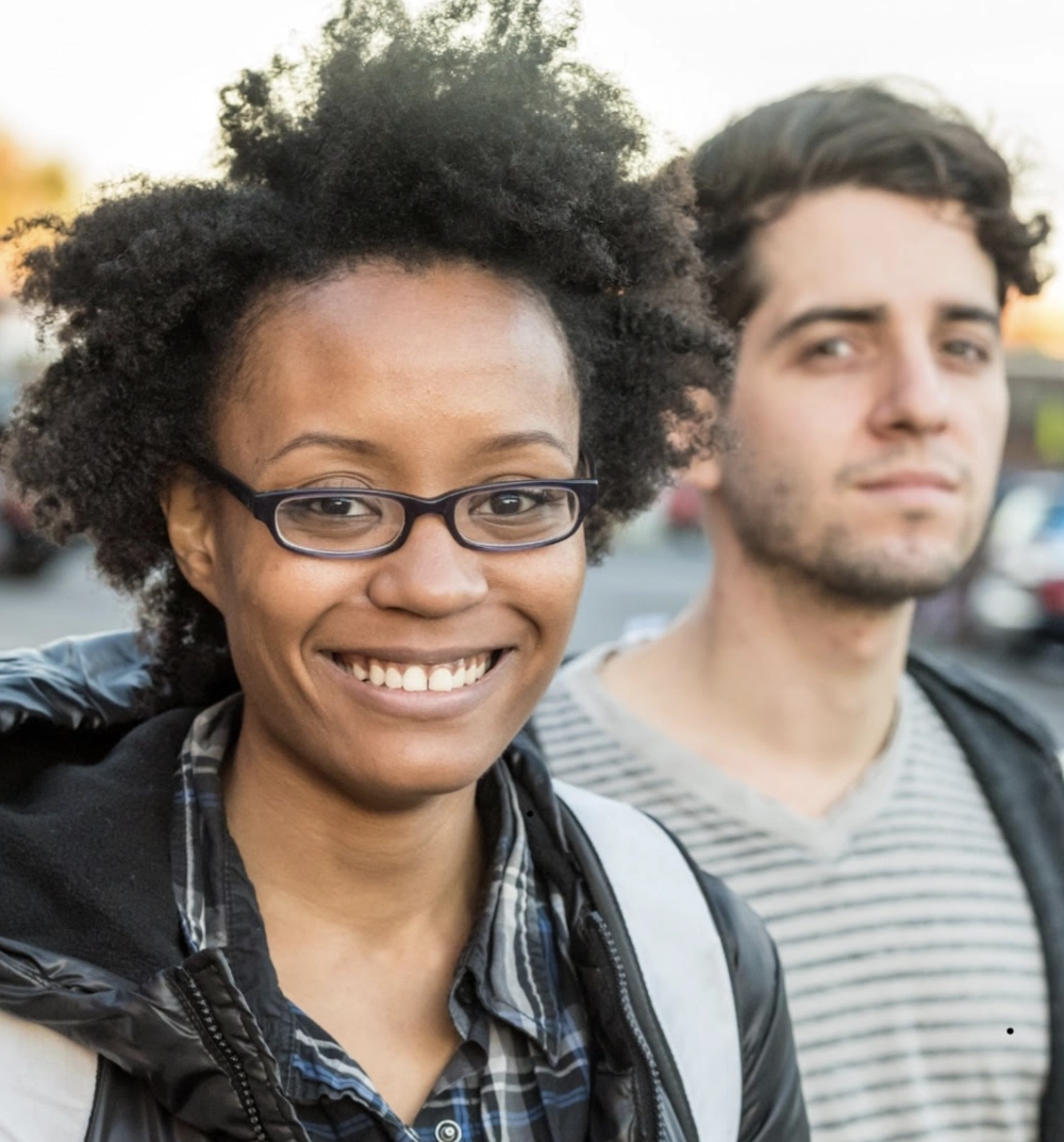 Smiling woman with glasses and curly hair in a plaid shirt and jacket, standing outdoors with a man in a striped shirt behind her.