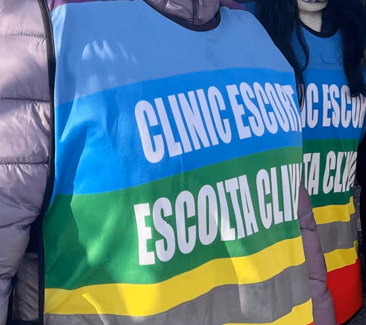 Close-up of two people wearing colorful vests with text 'CLINIC ESCORT' and its Spanish translation 'ESCOLTA CLINICA' in white letters.