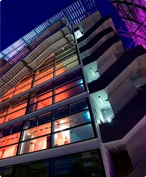Vista dal basso verso l'alto di un edificio moderno con ampie finestre illuminate da luci arancioni, blu e viola di sera.