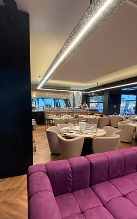 Interno elegante di un ristorante con divano viola in primo piano, tavoli apparecchiati e luci moderne sul soffitto.
